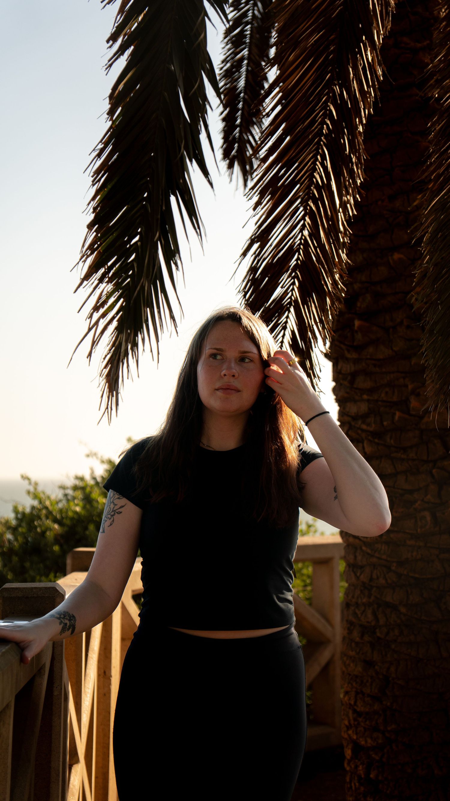 Sunset portrait under palm fronds showing someone in black attire leaning against a wooden railing.