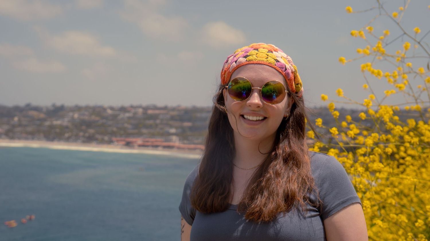 Person in colorful headscarf smiles with ocean waves and yellow flowers creating scenic backdrop.