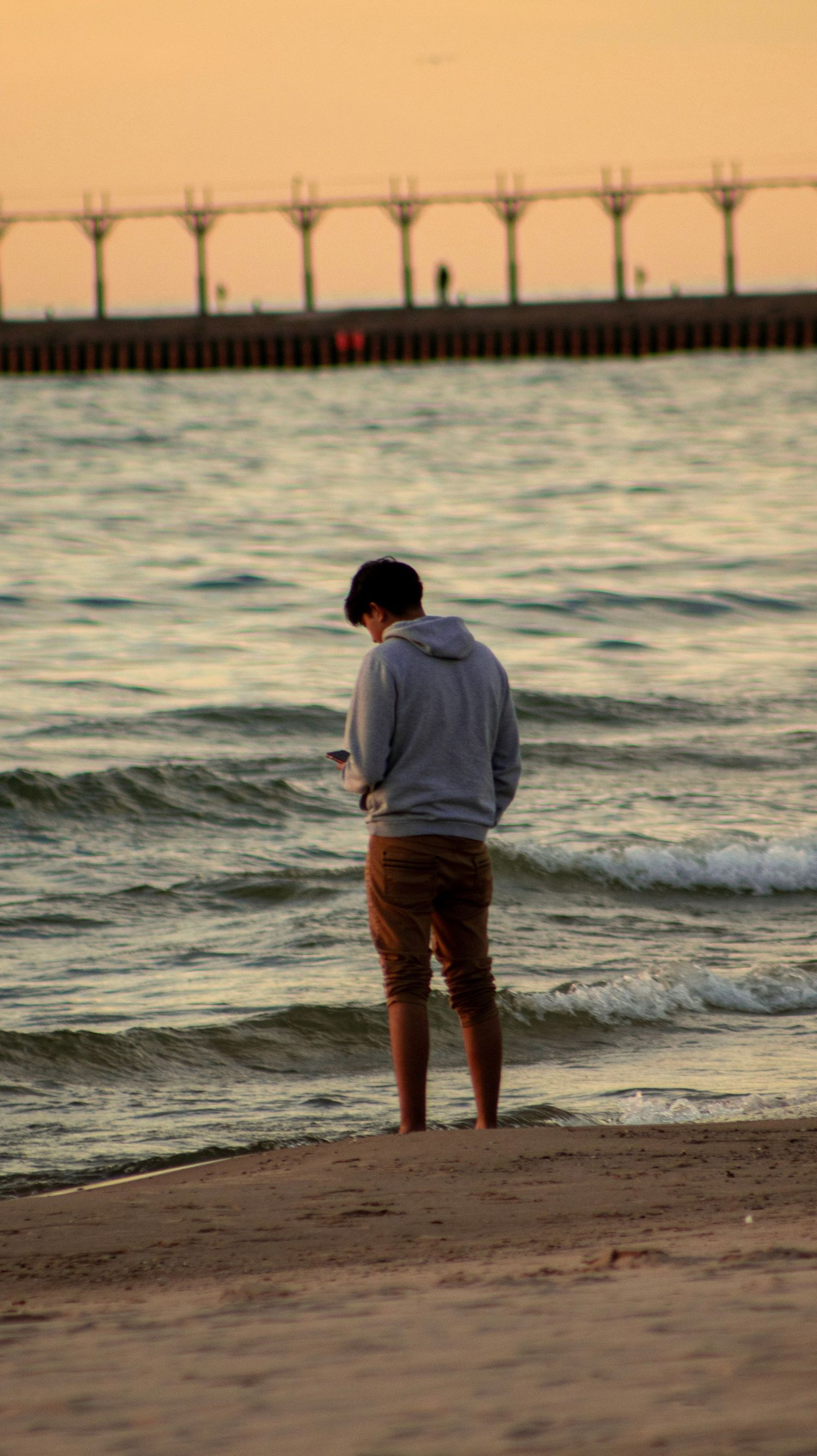 Person standing at beach edge watching waves near pier during golden sunset.
