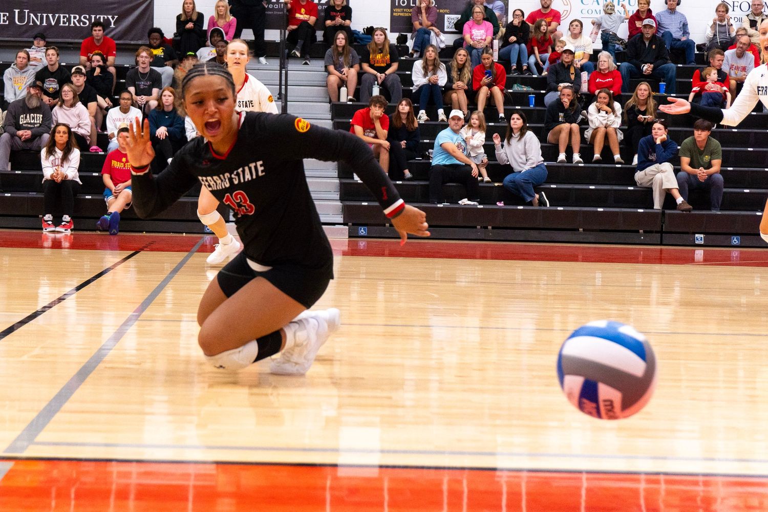 A volleyball player in a black jersey dives low on the court to dig a ball during a college match as spectators watch from the stands.