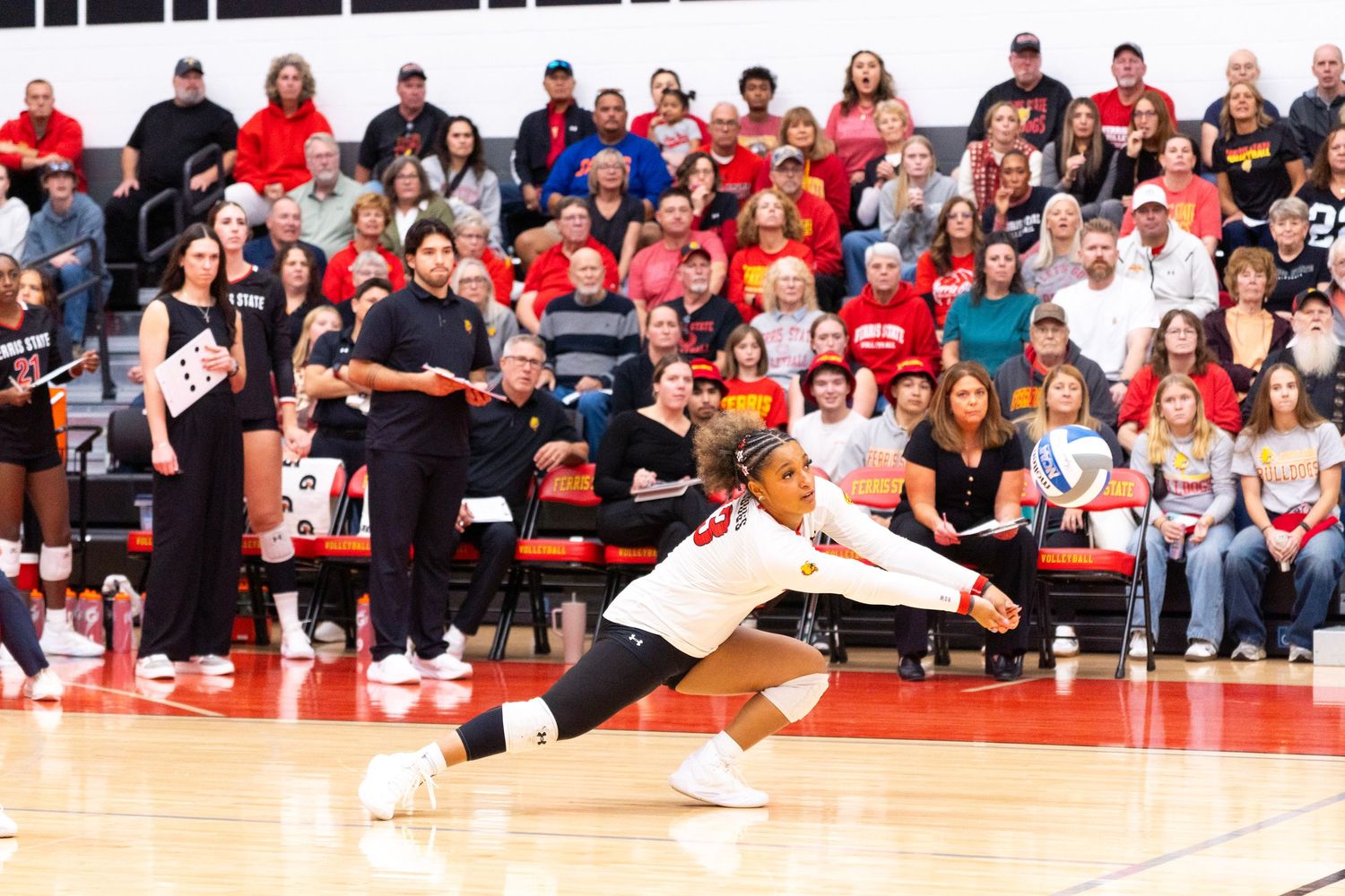 A volleyball player in a white jersey dives low to dig the ball during an indoor match while a crowd of fans in red watches from the stands.