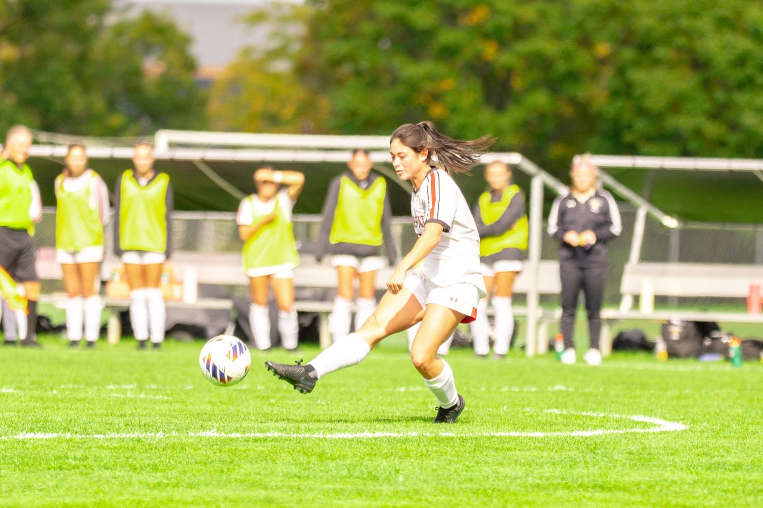 A female soccer player in a white uniform kicks the ball across a green field while teammates in neon yellow bibs watch from the sideline.