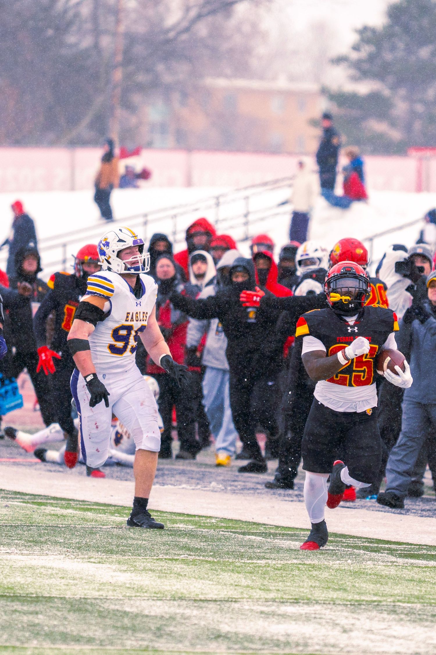 Football players compete in snowy conditions during a winter game with spectators watching from the sidelines.