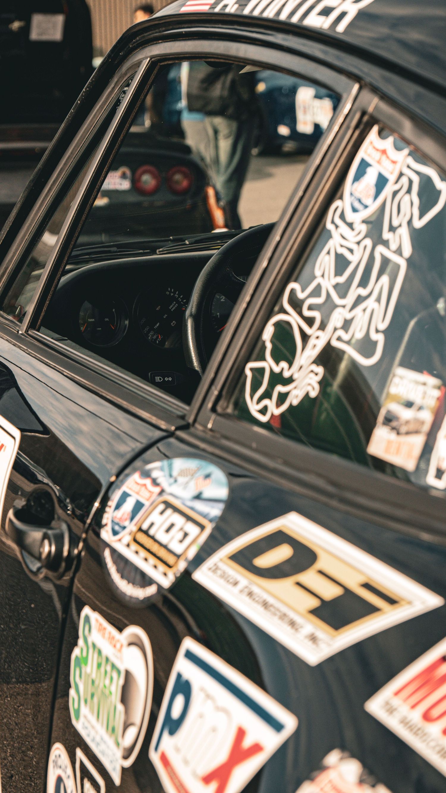Close-up of a black racing Porsche's door panel covered in various motorsport sponsor stickers and decals.