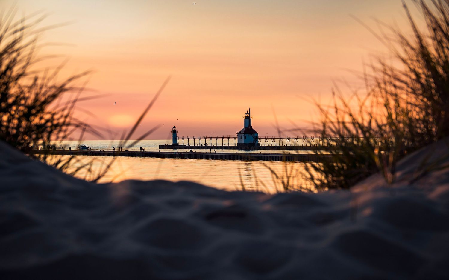Dramatic silhouette of a lighthouse and pier against a warm sunset sky viewed through coastal grasses.