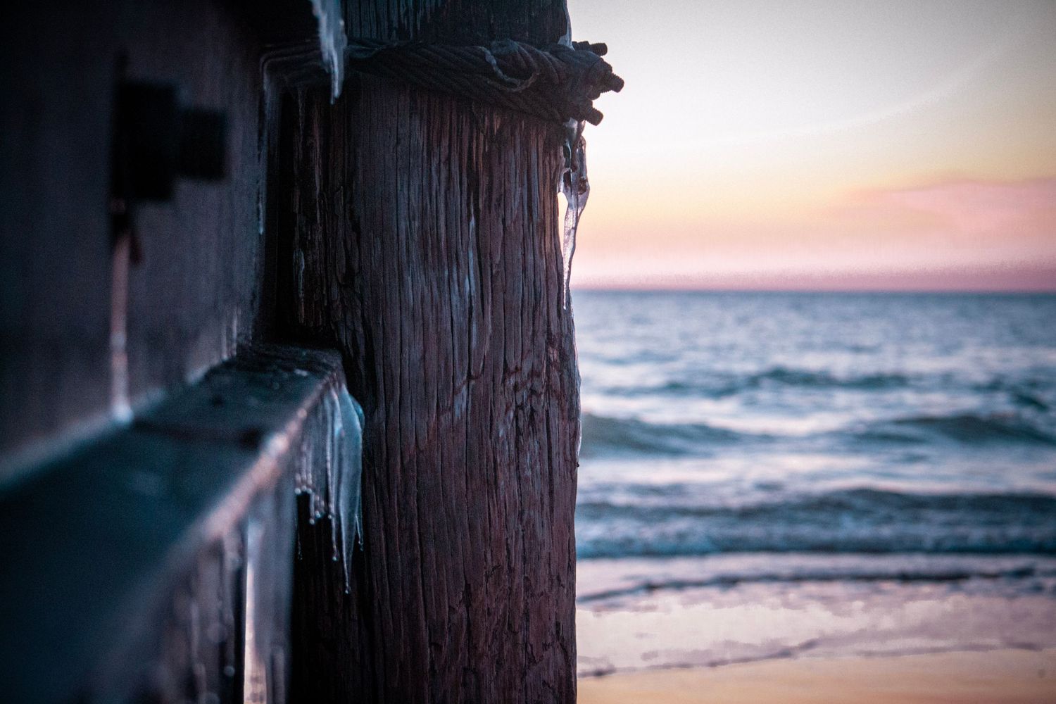 Wooden post with ice formations at sunset on a beach with ocean waves in the background.