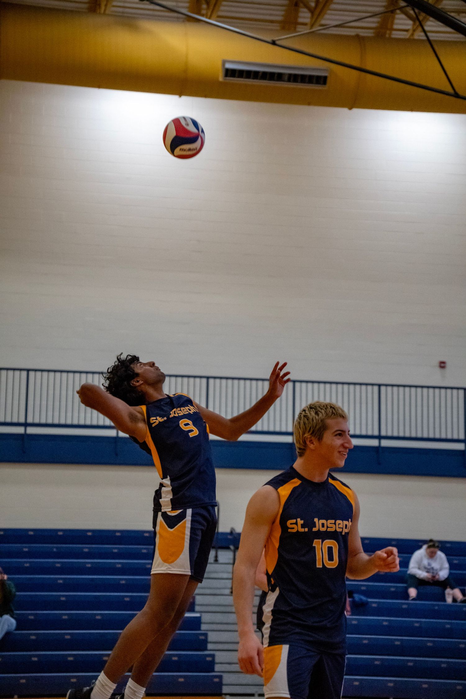 Volleyball players compete at the net as the ball floats above during an indoor match.