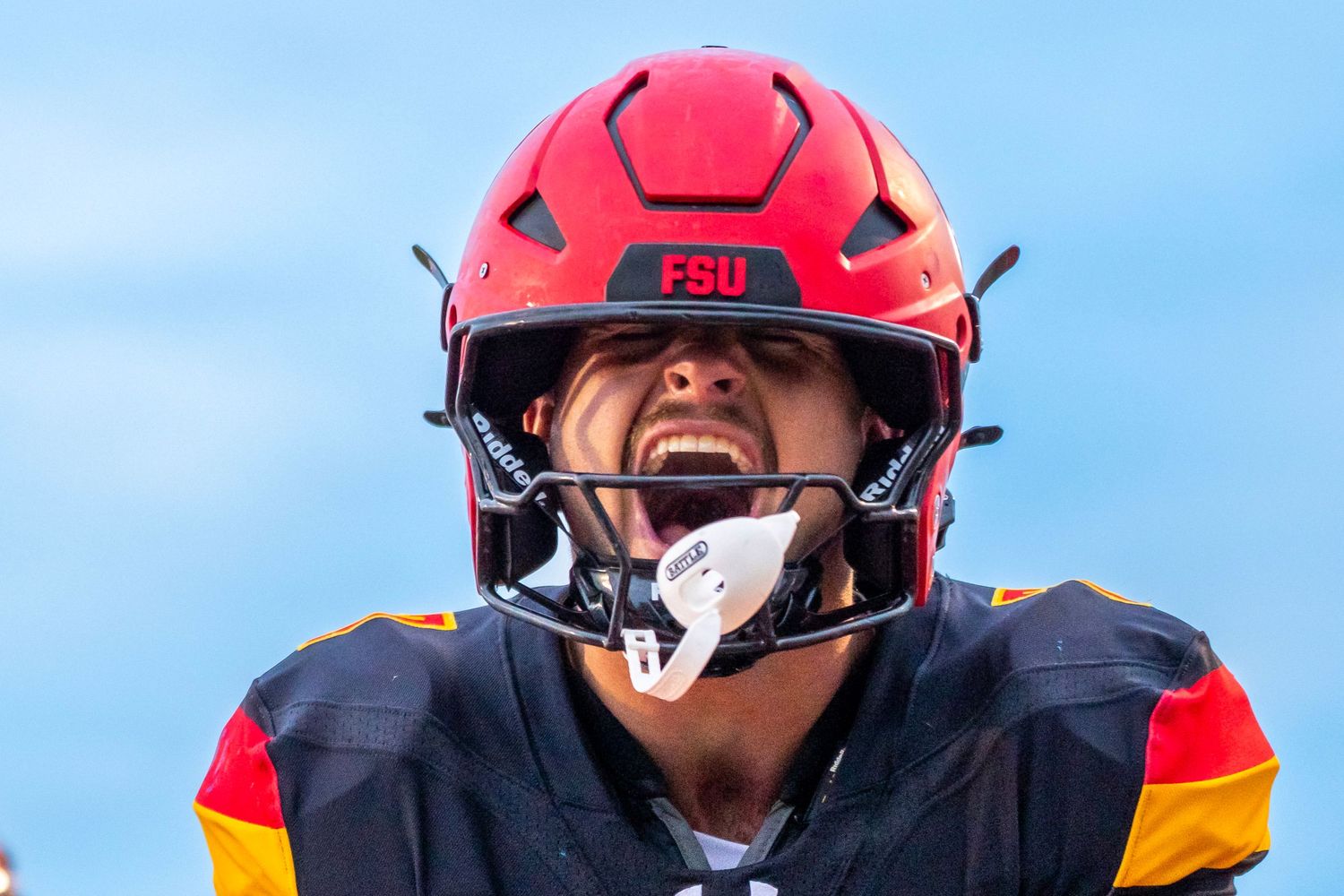 Football player in a red FSU helmet and black jersey shouts with intensity, mouthguard hanging from his mouth, against a clear blue sky.