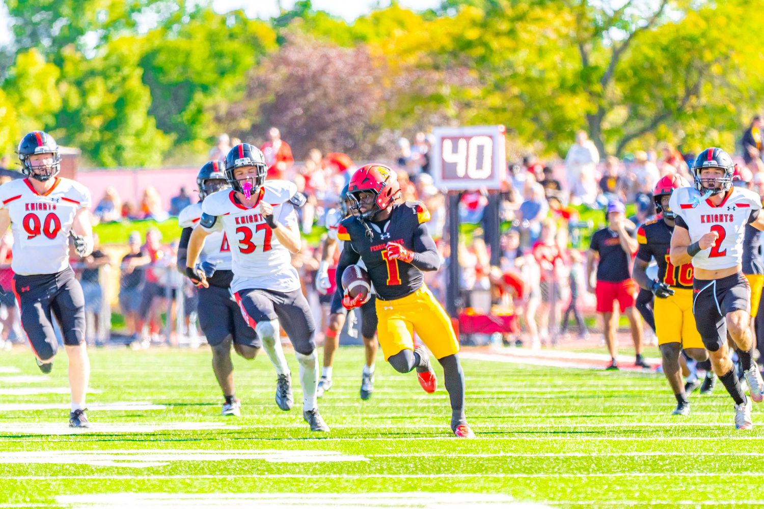 Football ball carrier in a black and yellow uniform sprints downfield past defenders in white jerseys during a college game near the 40-yard