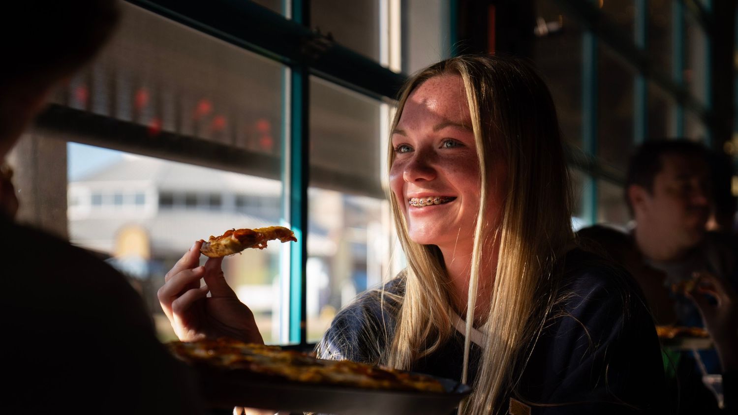 Woman with long blonde hair laughs while holding a sandwich in a dimly lit restaurant setting.
