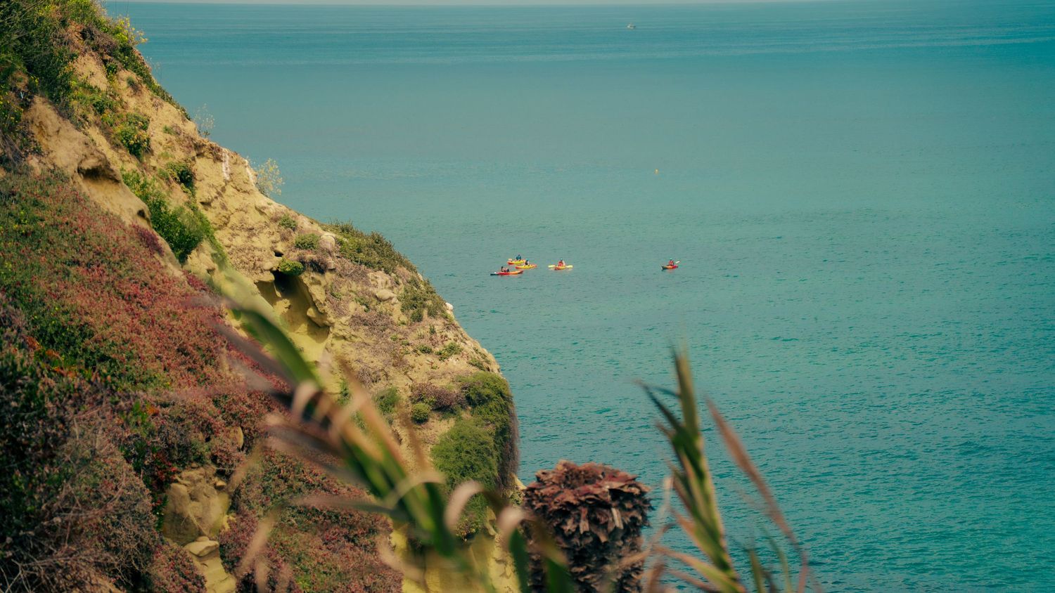 Orange kayakers dot the turquoise ocean waters near steep coastal cliffs.