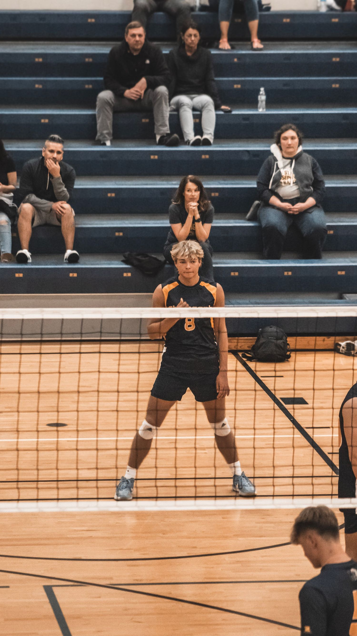 Player stands ready at net during indoor volleyball game with spectators seated in bleachers.