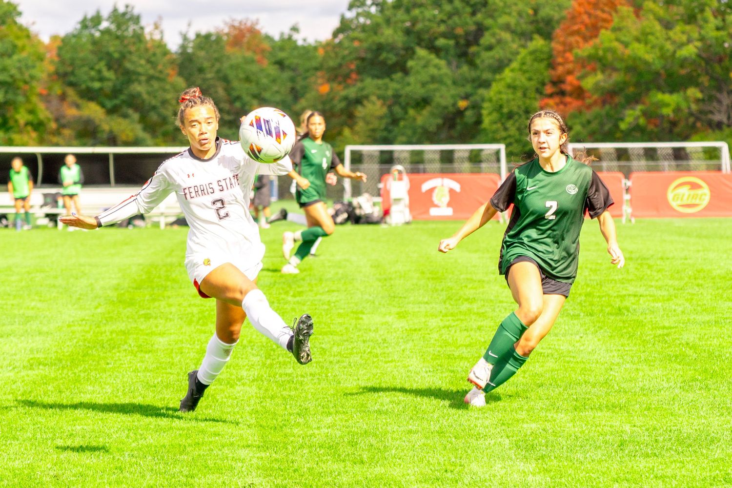 Women's soccer player in a white jersey kicks the ball past a defender in green during a match on a sunny grass field.