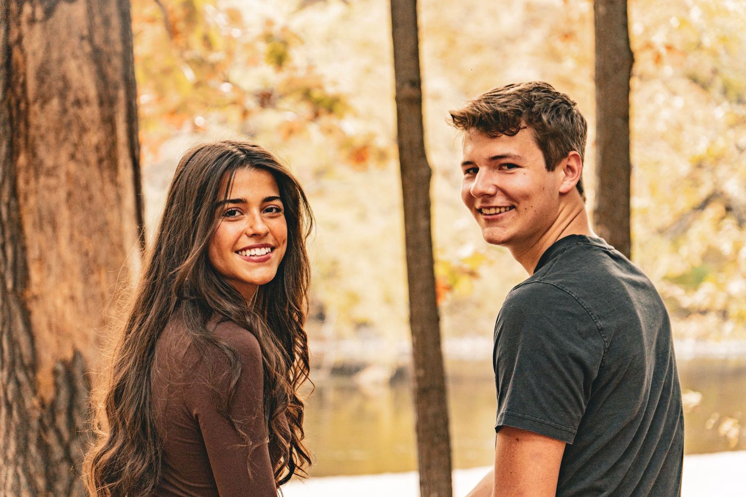 A smiling young woman with long dark hair and a young man in a gray t-shirt look back at the camera among autumn trees beside a lake.