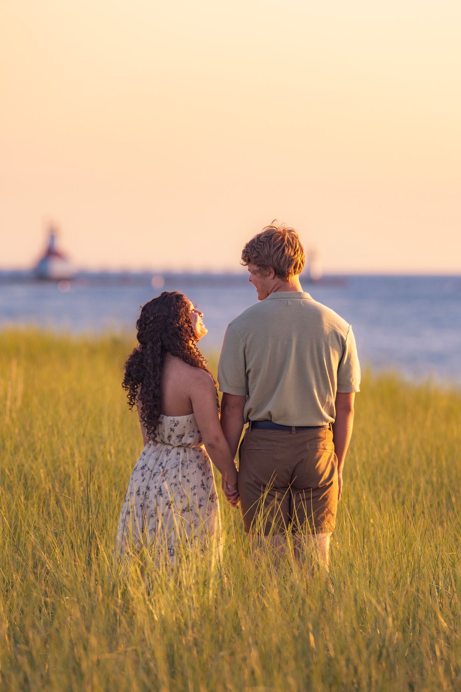 A couple stands together in a golden seaside meadow at sunset with a lighthouse visible in the distance.