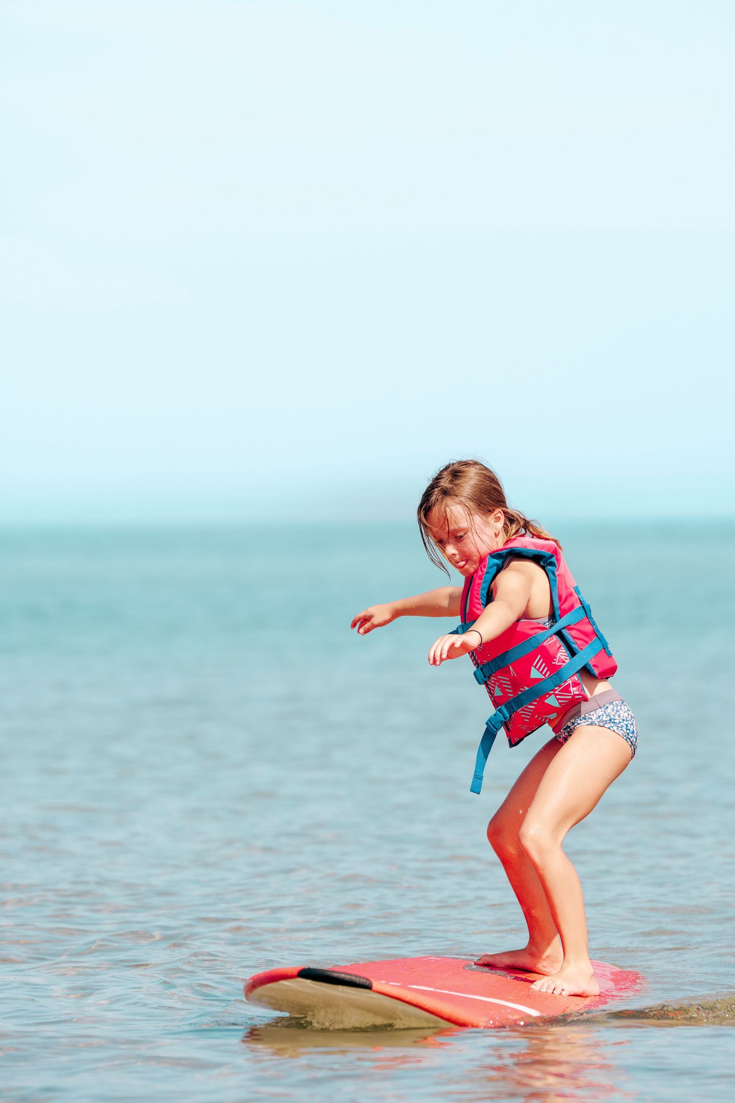 A child practices surfing on a pink board in turquoise ocean waters while wearing a red life vest.