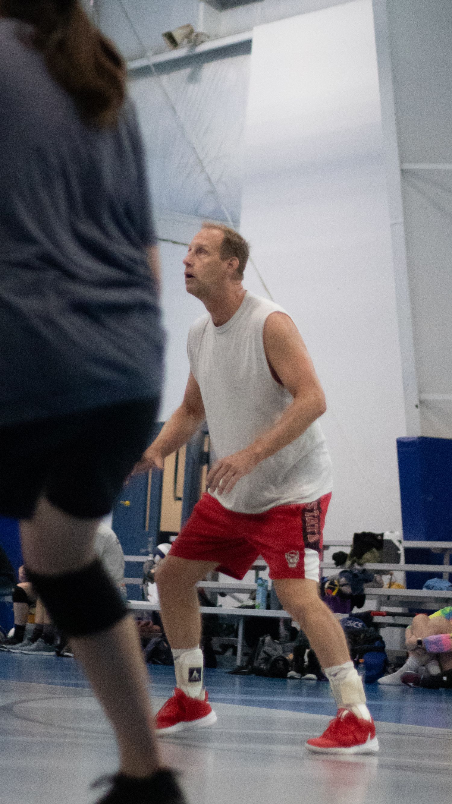 Indoor volleyball player in white tank top and red shorts preparing to receive during a match.