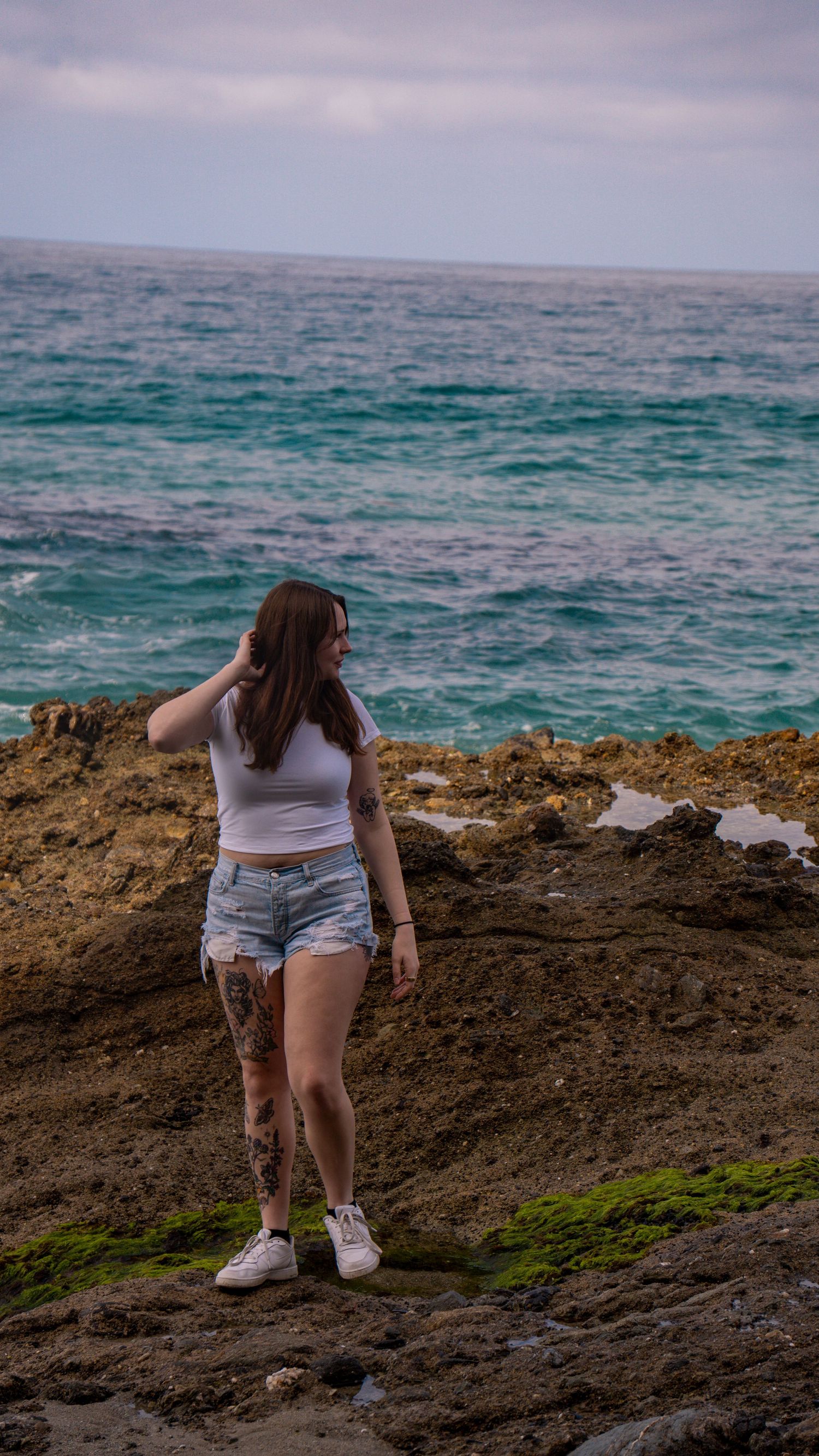 Figure standing on dark rocky shore against turquoise ocean backdrop.