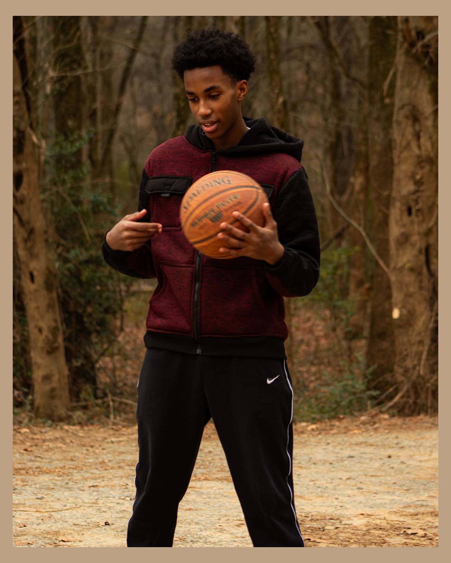 Person in burgundy vest holding basketball while standing in front of bare trees.