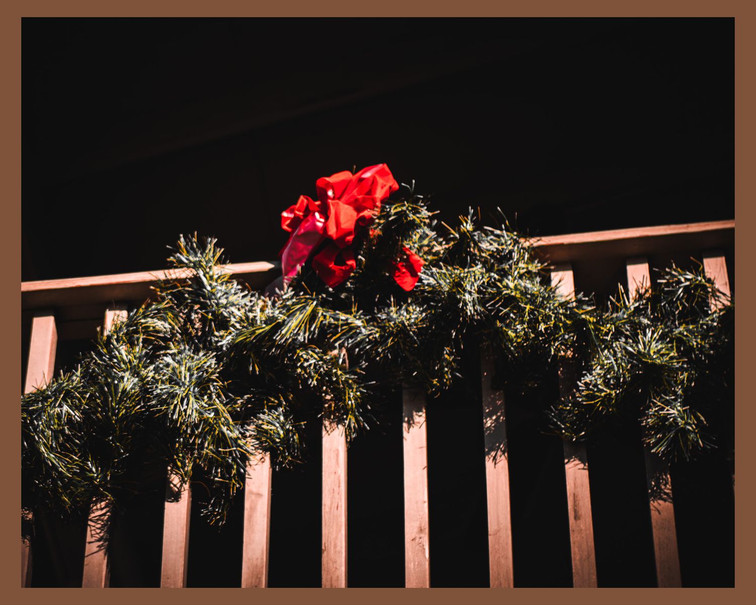 Red flowers and evergreen garland decorating wooden railing against dark background.