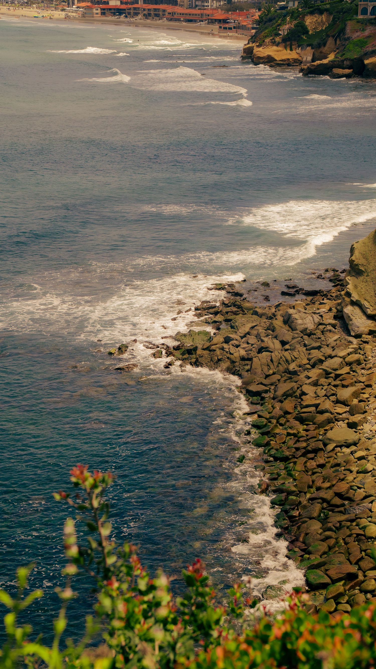 Rocky coastline with crashing waves against jagged cliffs and beach homes in the distance.