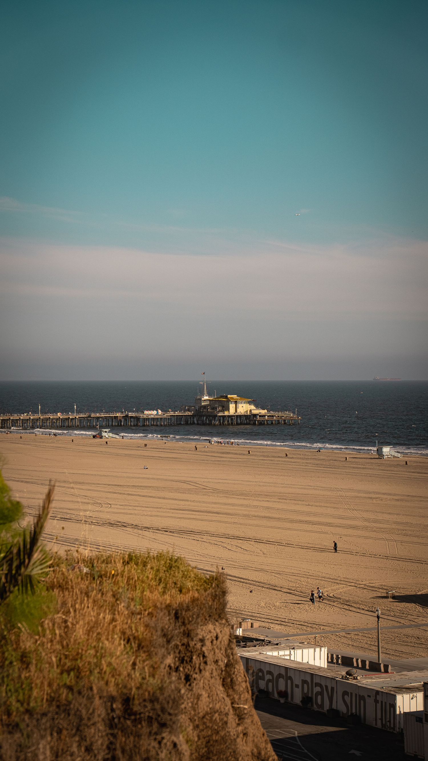 Wide sandy beach stretches along the coast with a pier visible in the distance under a turquoise sky.