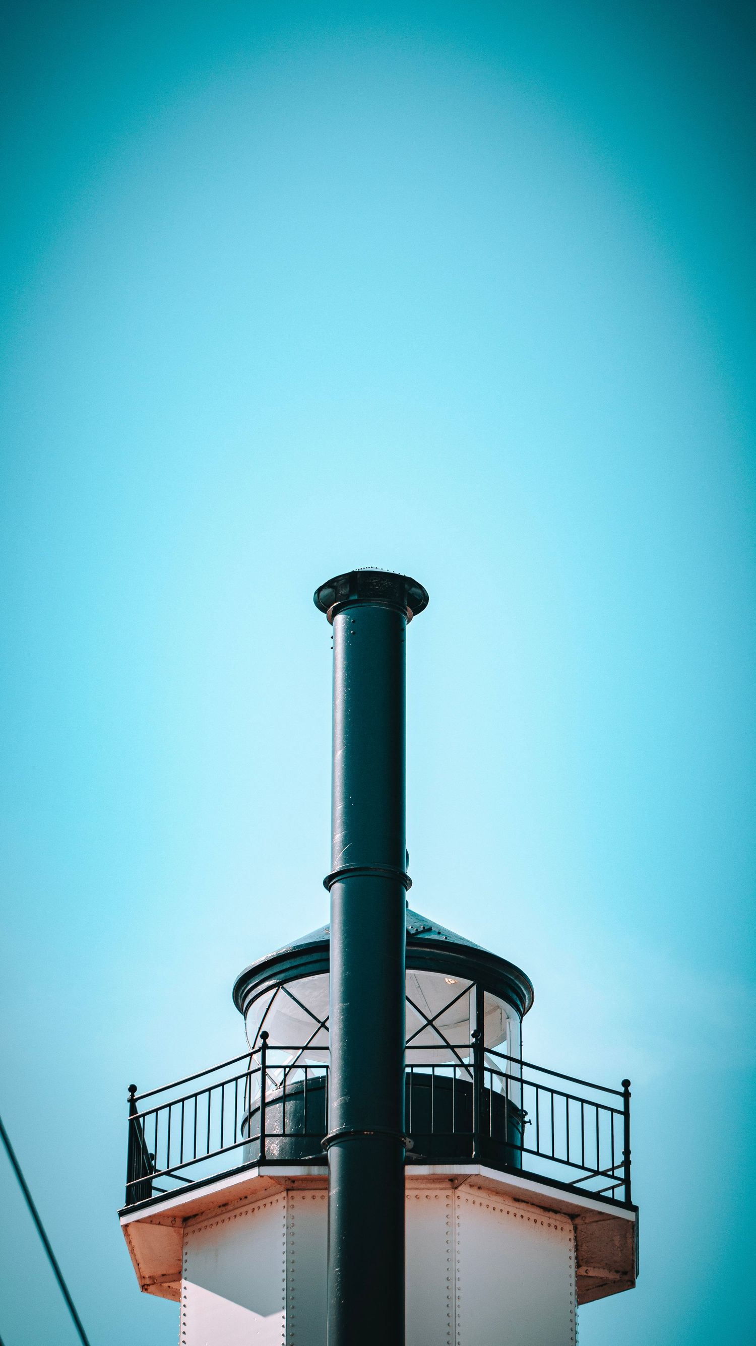 Minimalist architectural detail of lighthouse tower with metal railing against bright blue sky.