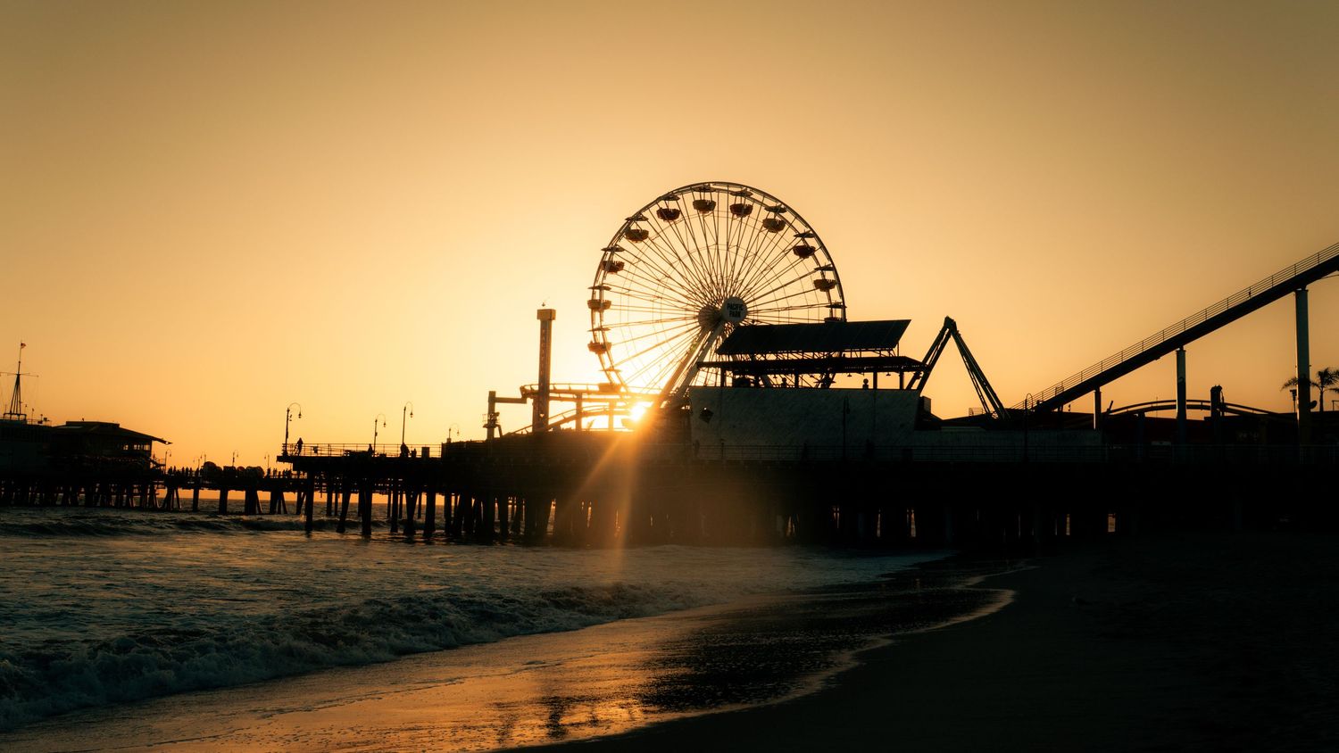 Golden sunset silhouetting pier and Ferris wheel with dramatic sky.