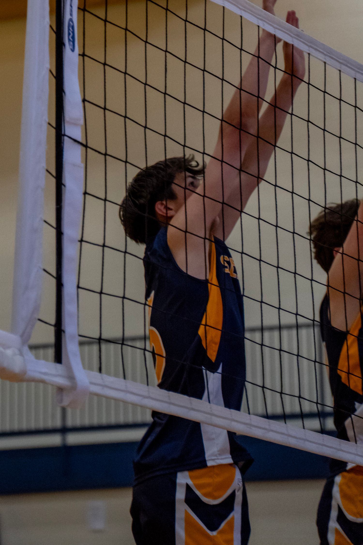Players reaching up to block at volleyball net during indoor match.