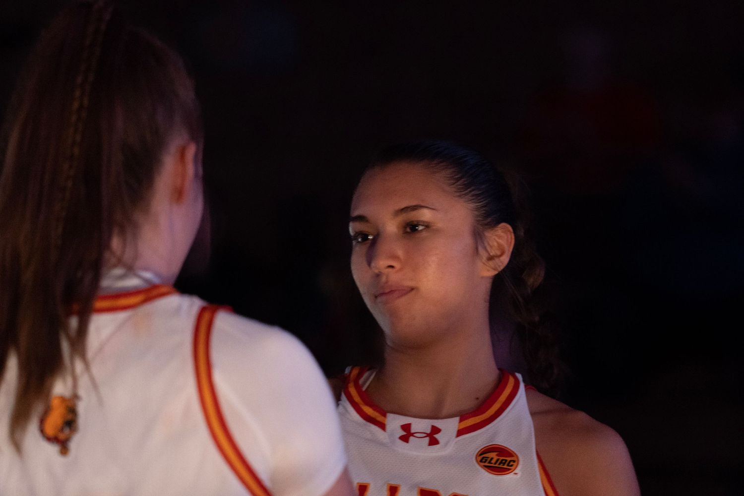 Two basketball players in orange and white Under Armour uniforms stand together during a dark indoor game.