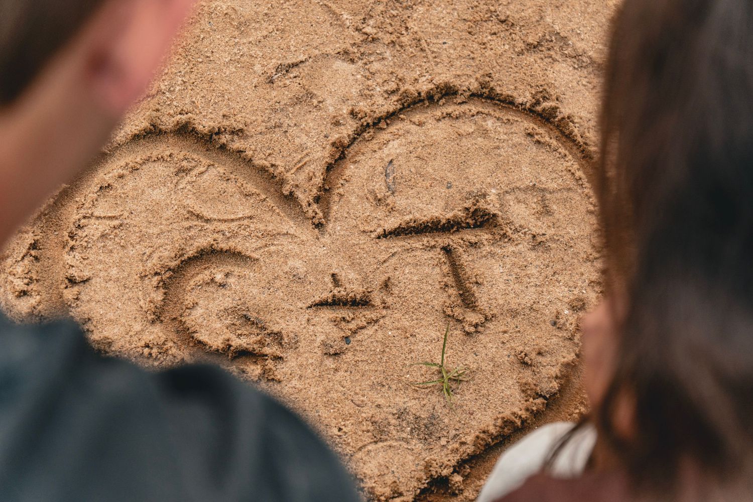 Two people look down at a heart drawn in smooth sand, with the outline and small marks carved into the beach surface.