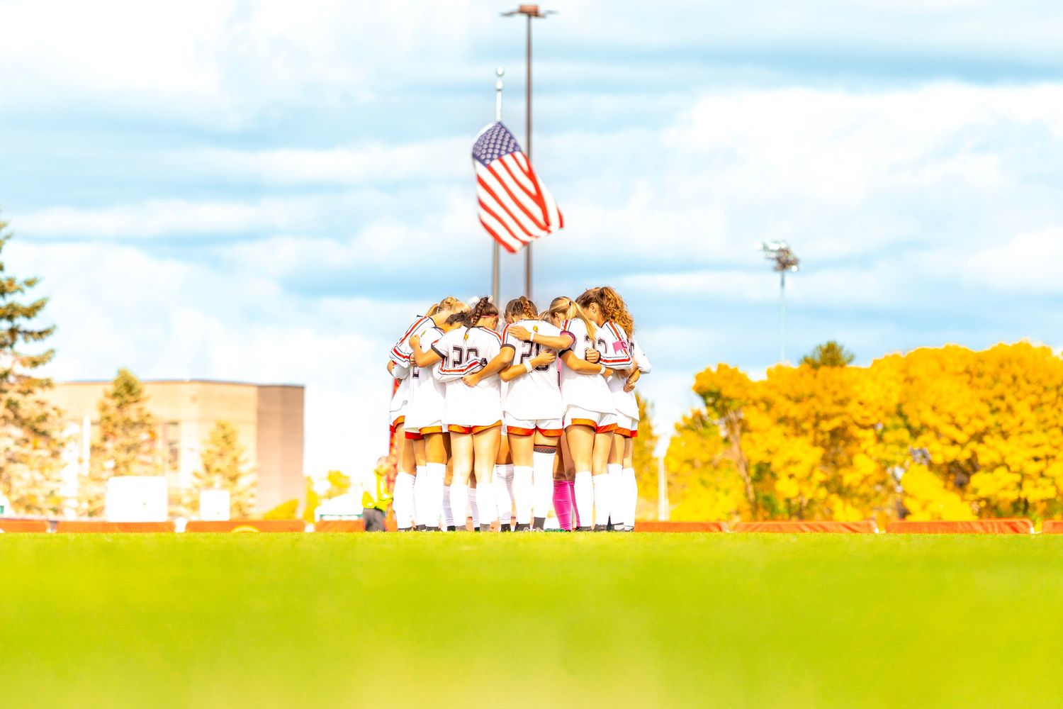 Soccer team in white uniforms huddles together on a green field beneath an American flag, with golden autumn trees and a building in the bac