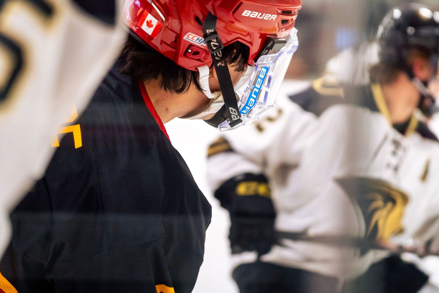 Hockey player in a red Bauer helmet with visor faces an opponent in a white and black jersey during a close-up faceoff on the ice.
