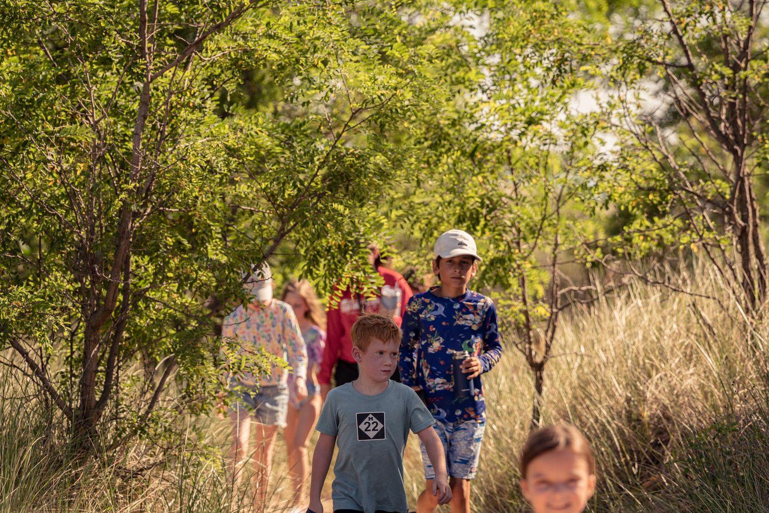 Group of hikers walking along a sunny nature trail surrounded by green foliage and tall grass.