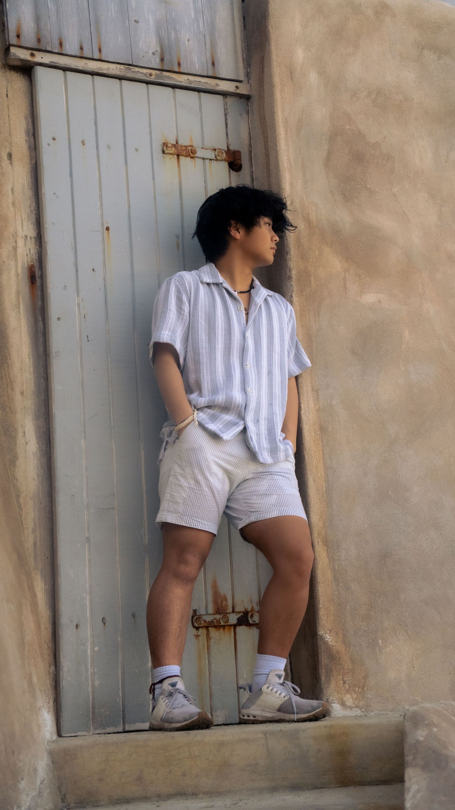 Figure in striped shirt and white shorts leaning against rustic door in urban setting.