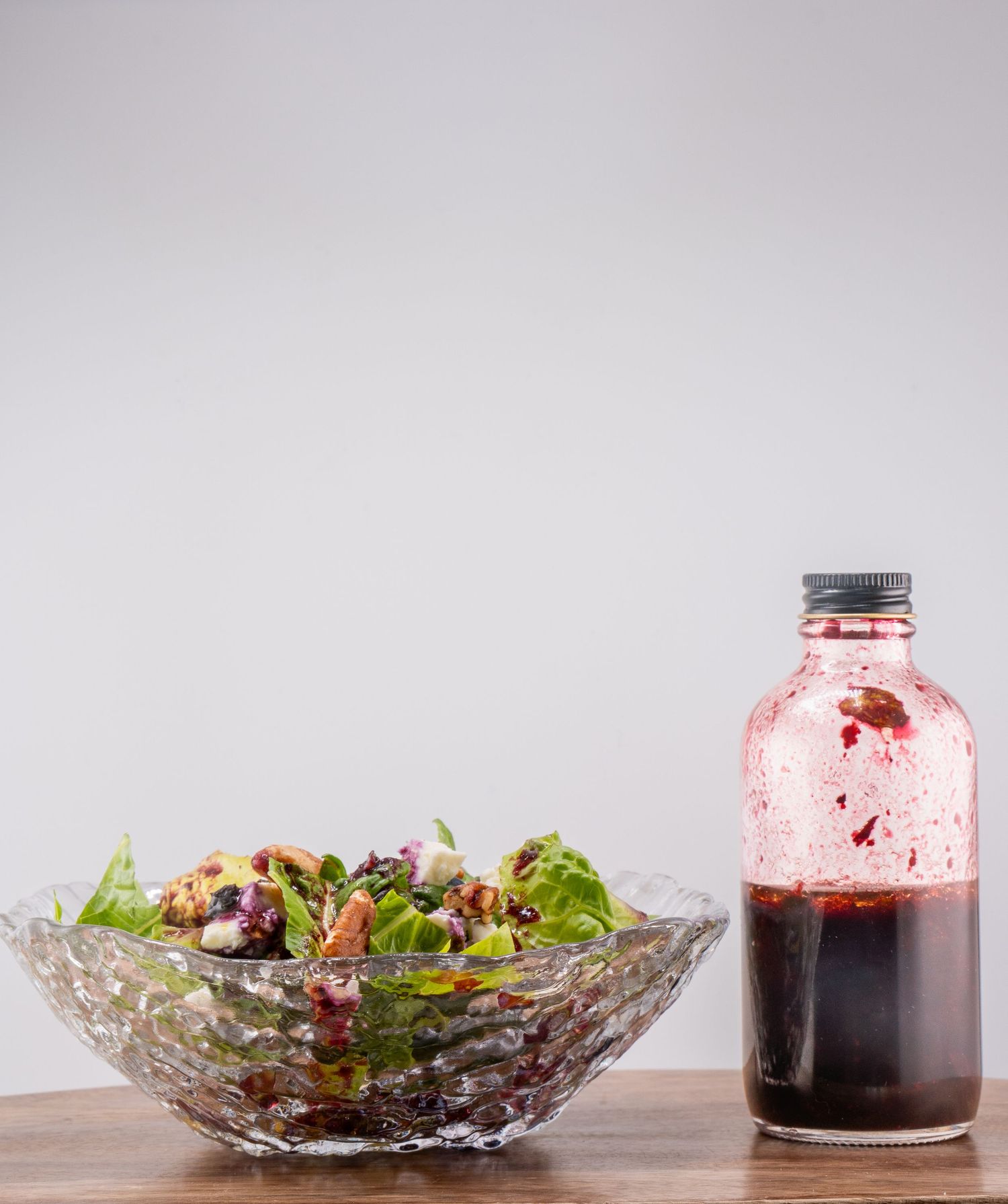 A glass jar of red berry syrup and salad ingredients arranged on a wooden surface.