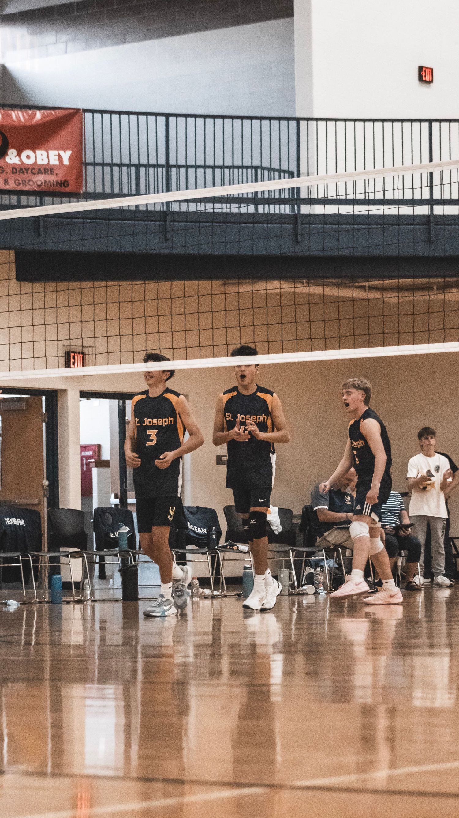 Three volleyball players in black St. Joseph uniforms stand on court during game break.