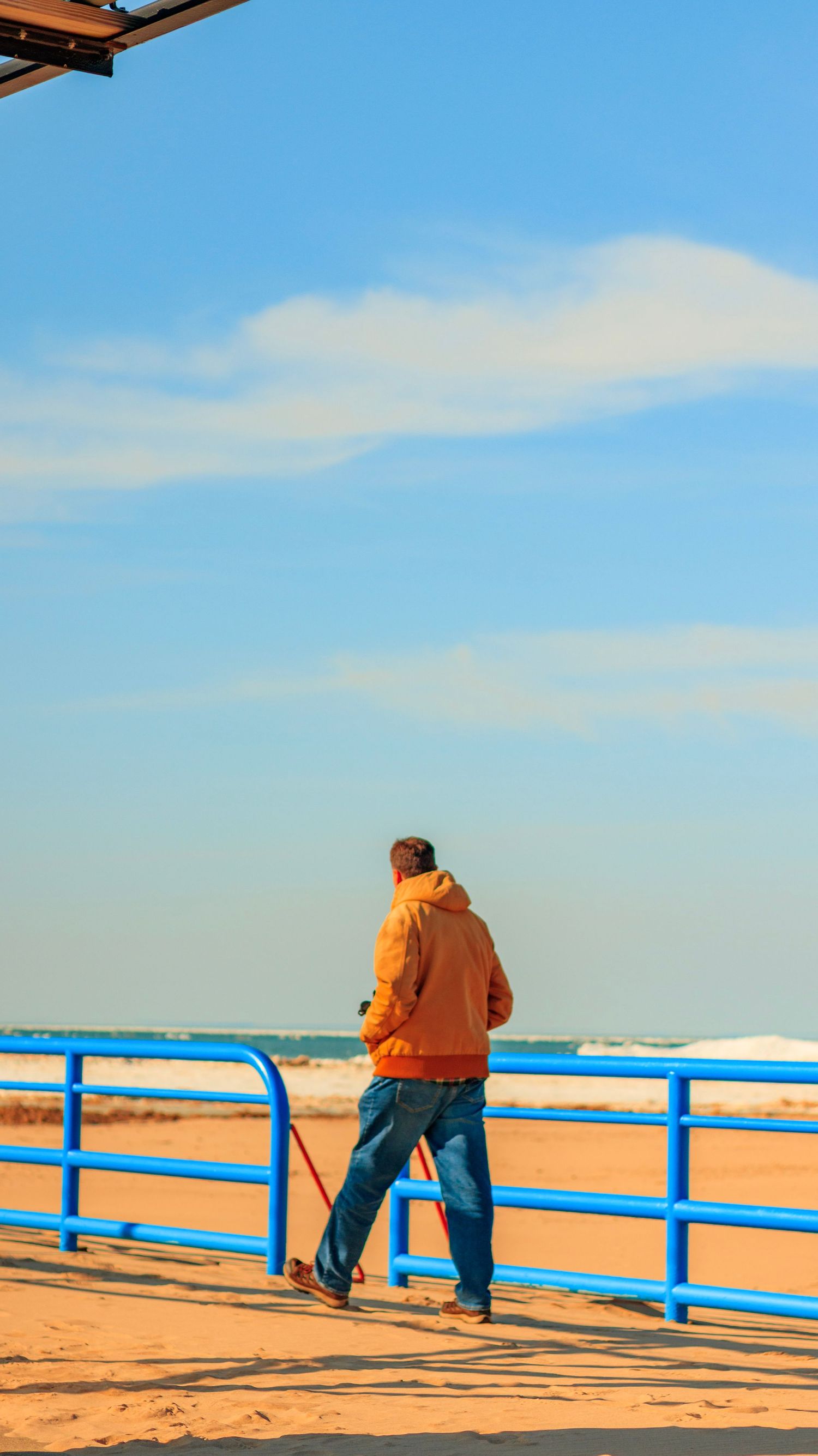 A figure in brown clothing leans on a bright blue railing overlooking a beach at sunset.