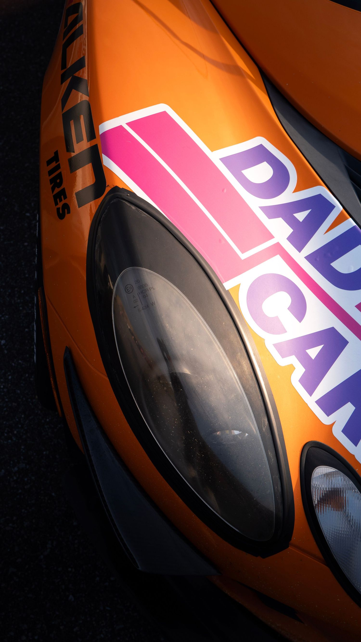 Close-up of orange race car headlight with DADCA racing decal visible.