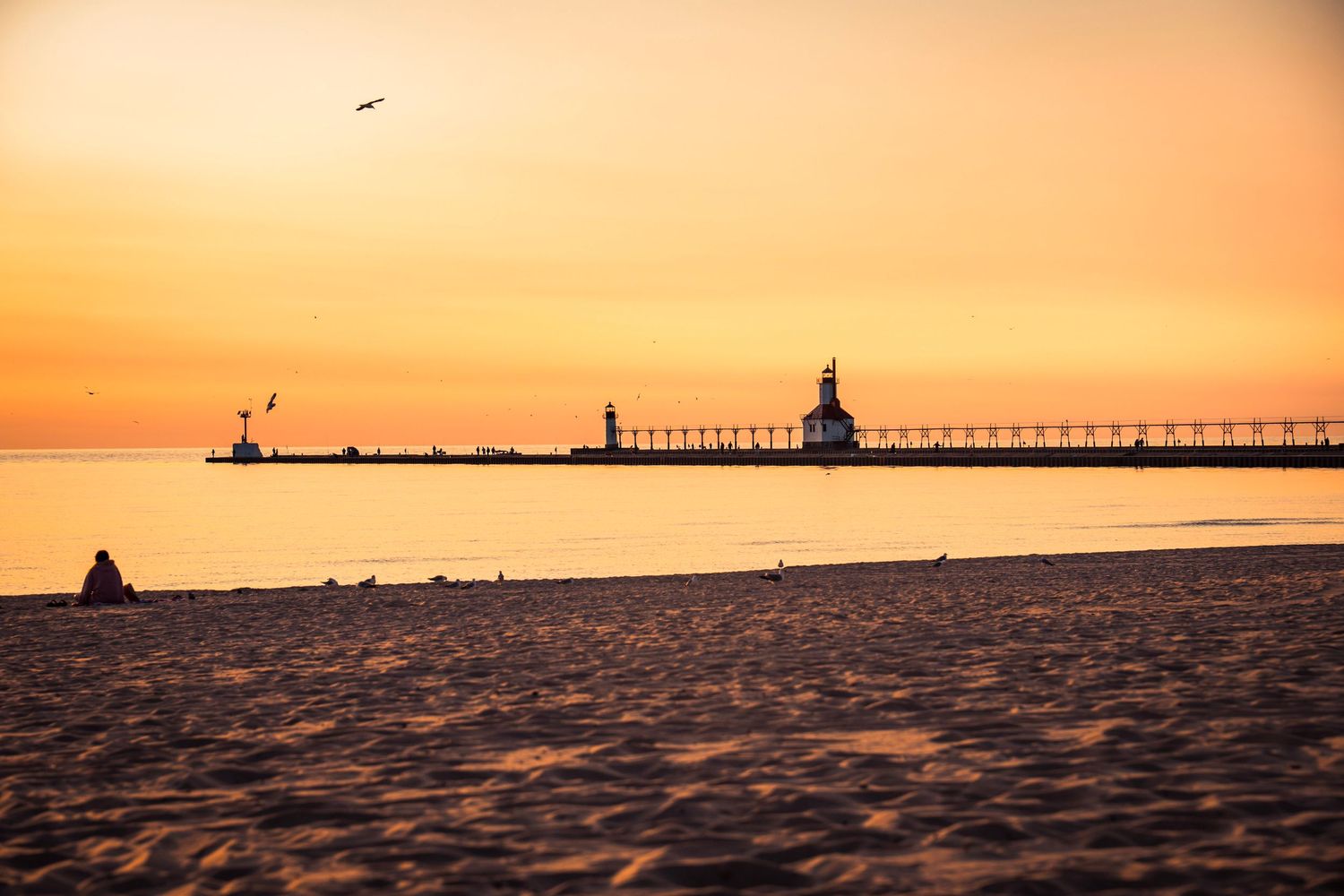 Sandy beach stretching towards a distant lighthouse and pier during a vibrant orange sunset.