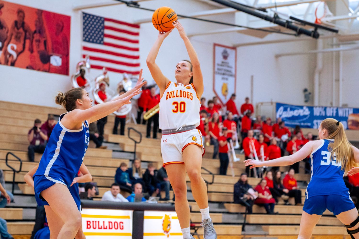 Women's basketball player in a white and orange number 30 jersey shoots over two defenders in blue uniforms as fans watch from the stands.