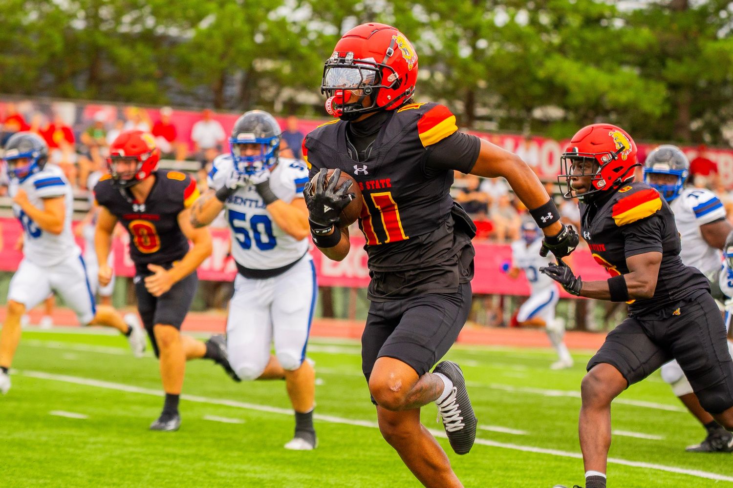 Football player in black and orange uniform and red helmet runs with the ball down the field as teammates and white-jersey opponents chase.