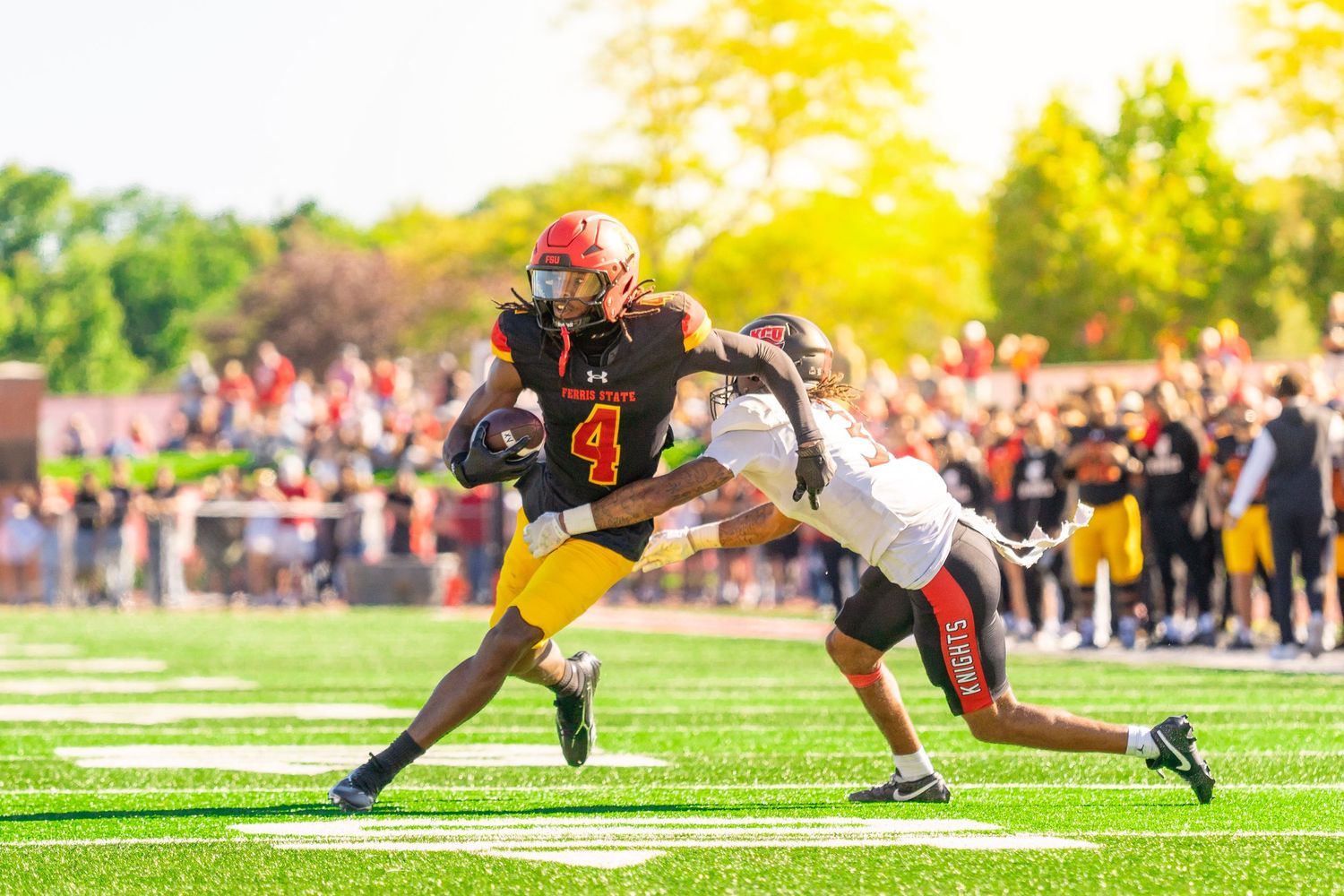 Football player in red helmet and black jersey number 4 carries the ball and stiff-arms a defender during a college game.