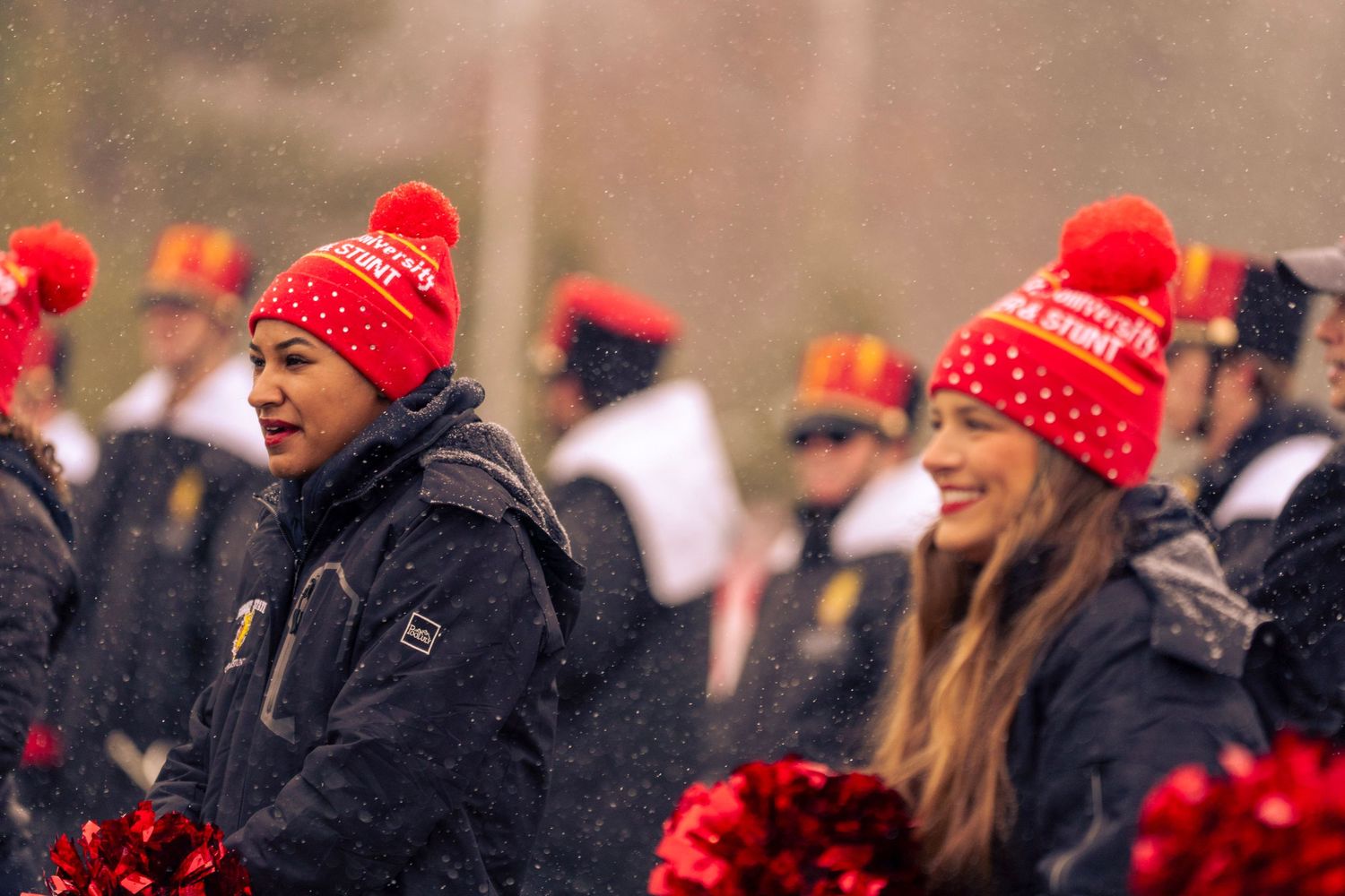 Spectators wearing matching red winter hats watch a sporting event on a cold day with cheerleaders visible in background.