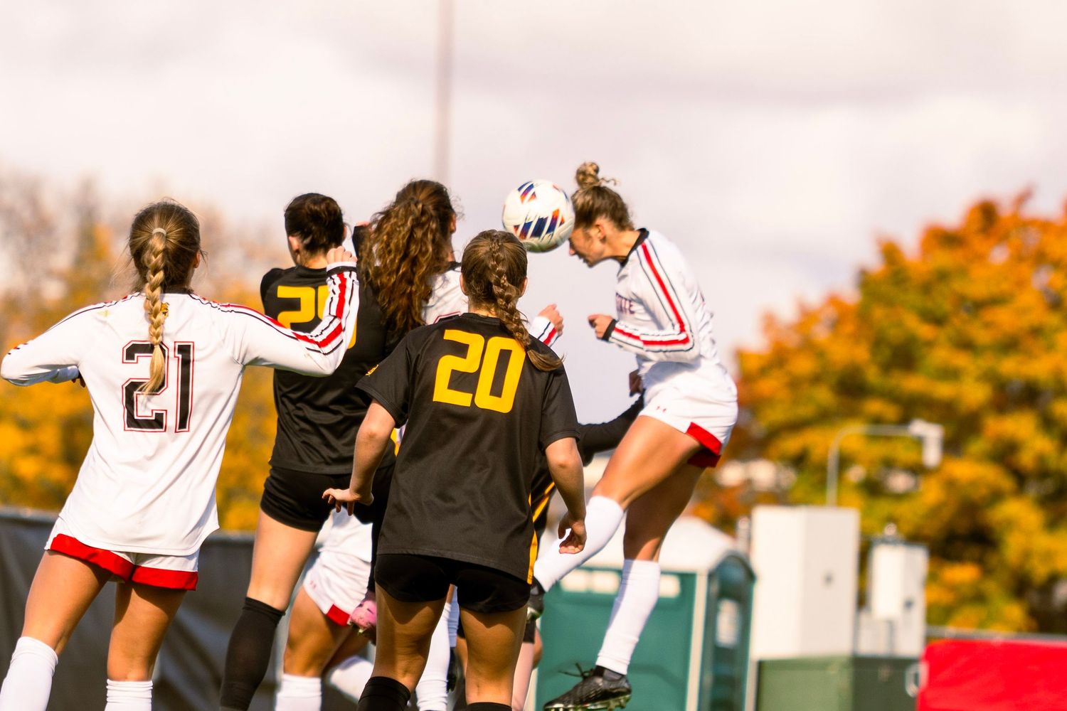 Soccer players in white and black jerseys compete for the ball during an intense match with autumn trees in background.