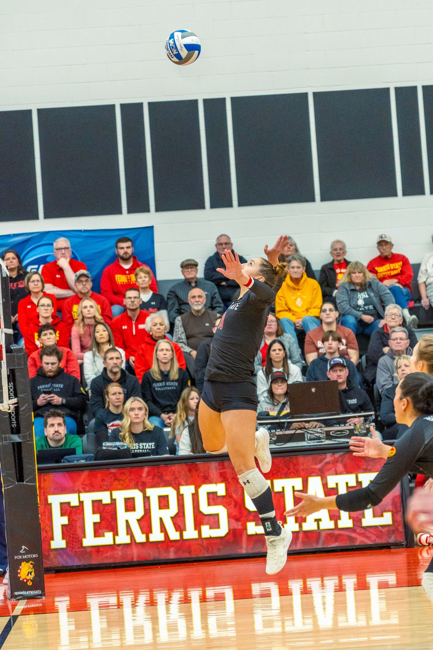Volleyball players leap at the net during a match with Ferris State banners and crowd visible in background.