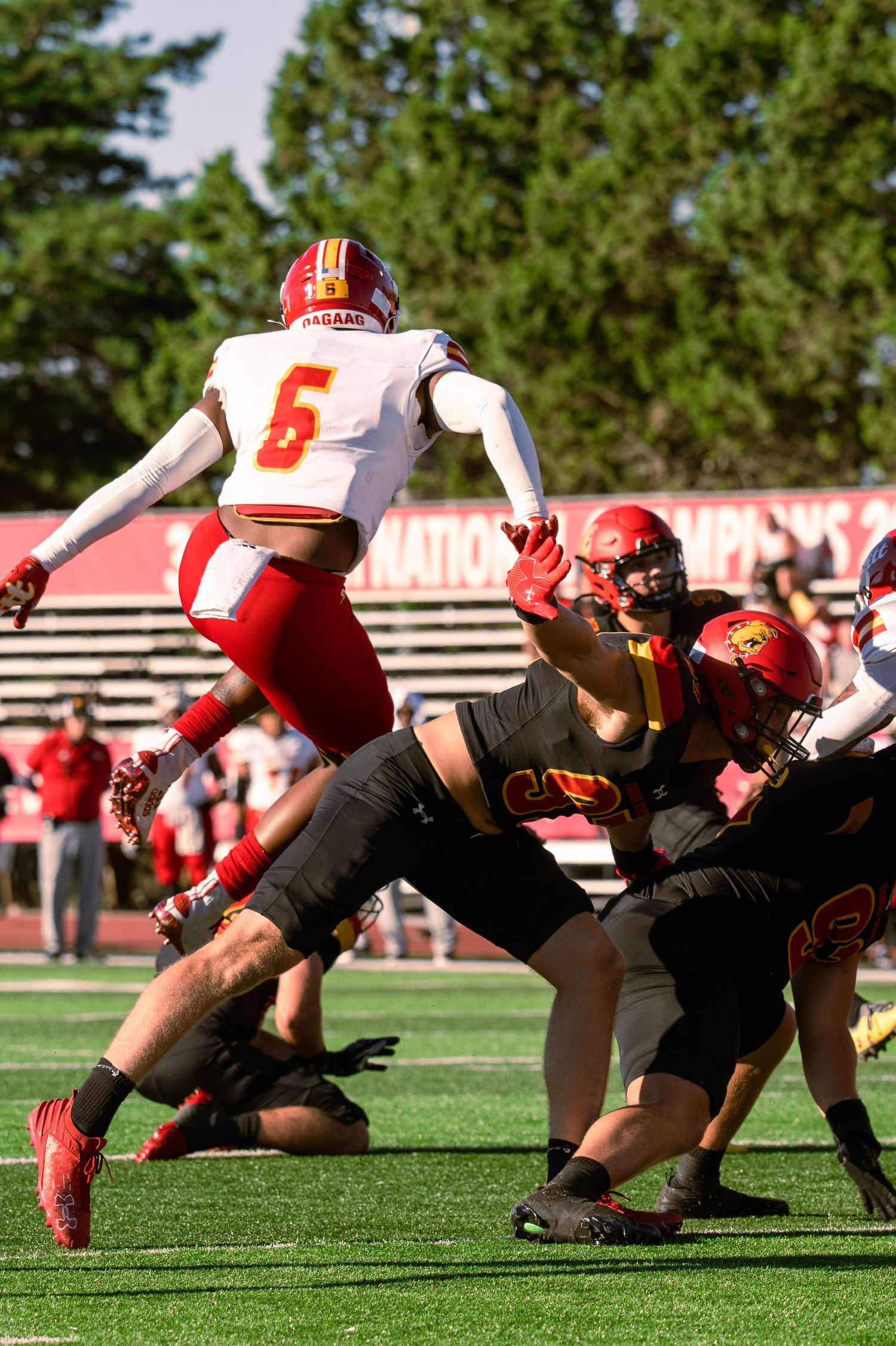 A football player in red jersey number 6 jumps over offensive linemen during a game on a sunny day.