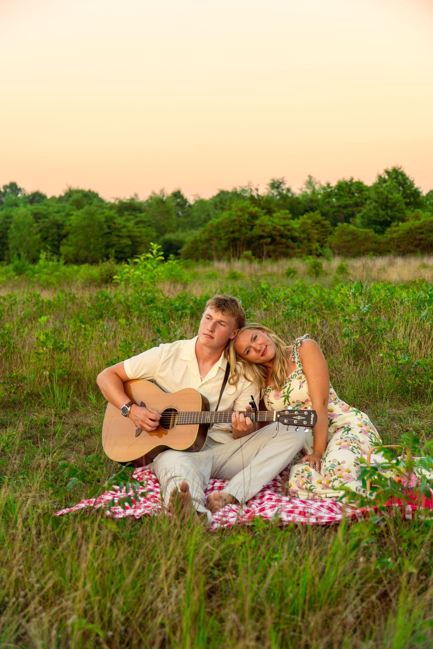 Young man plays acoustic guitar on a red checkered blanket while a woman rests her head on his shoulder in a grassy field at sunset.