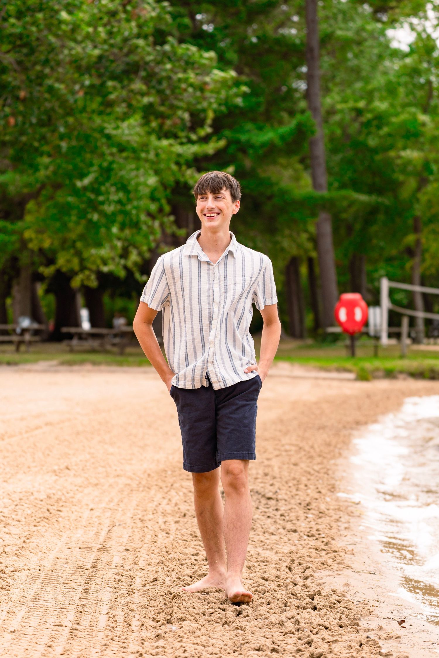 Smiling teen in a striped button-up shirt and navy shorts walks barefoot along a sandy lakeside beach with tall green trees behind him.