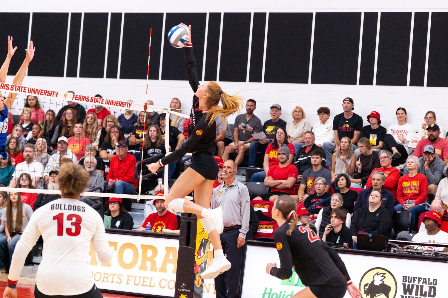 A volleyball player in a black uniform leaps high at the net to spike the ball while a teammate in white number 13 watches from the court.